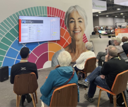 Attendees seated at the FamilyTreeDNA booth, listening to a presentation about DNA matching tools, with an interactive screen displaying genealogical insights.