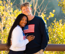 Olympic and Paralympic athletes Tara Davis-Woodhall and Hunter Woodhall smile together outdoors, wearing patriotic sweaters, ahead of their inspiring keynote at RootsTech 2025.