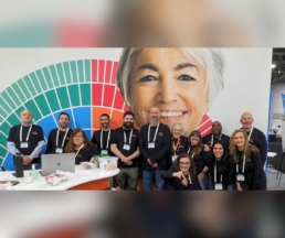The FamilyTreeDNA team at RootsTech 2025 poses in front of their vibrant booth, smiling and ready to welcome attendees. The backdrop features a large DNA-inspired design with a welcoming face, symbolizing the power of genetic genealogy in connecting family history.