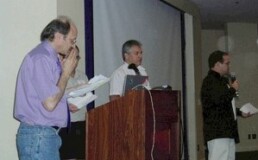 Bennett Greenspan speaks at a podium alongside Max Blankfeld and Matt Kaplan, with Bruce Walsh partially obscured, during the first International Conference on Genetic Genealogy put on by FamilyTreeDNA in 2004. Photo courtesy of ISOGG.