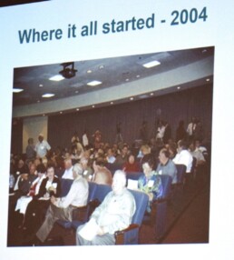 Slide projected during a later event showing an archival photo from the first International Conference on Genetic Genealogy (ICGG) in 2004, with attendees seated in a conference room. Text on the slide reads “Where it all started – 2004.”