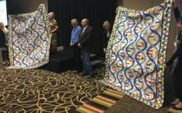 Roberta Estes presents handmade DNA-themed quilts to Bennett Greenspan and Max Blankfeld during the 2016 International Conference on Genetic Genealogy in Houston, Texas.